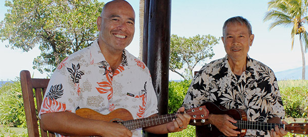 Two men smiling with ukuleles on the ocean side
